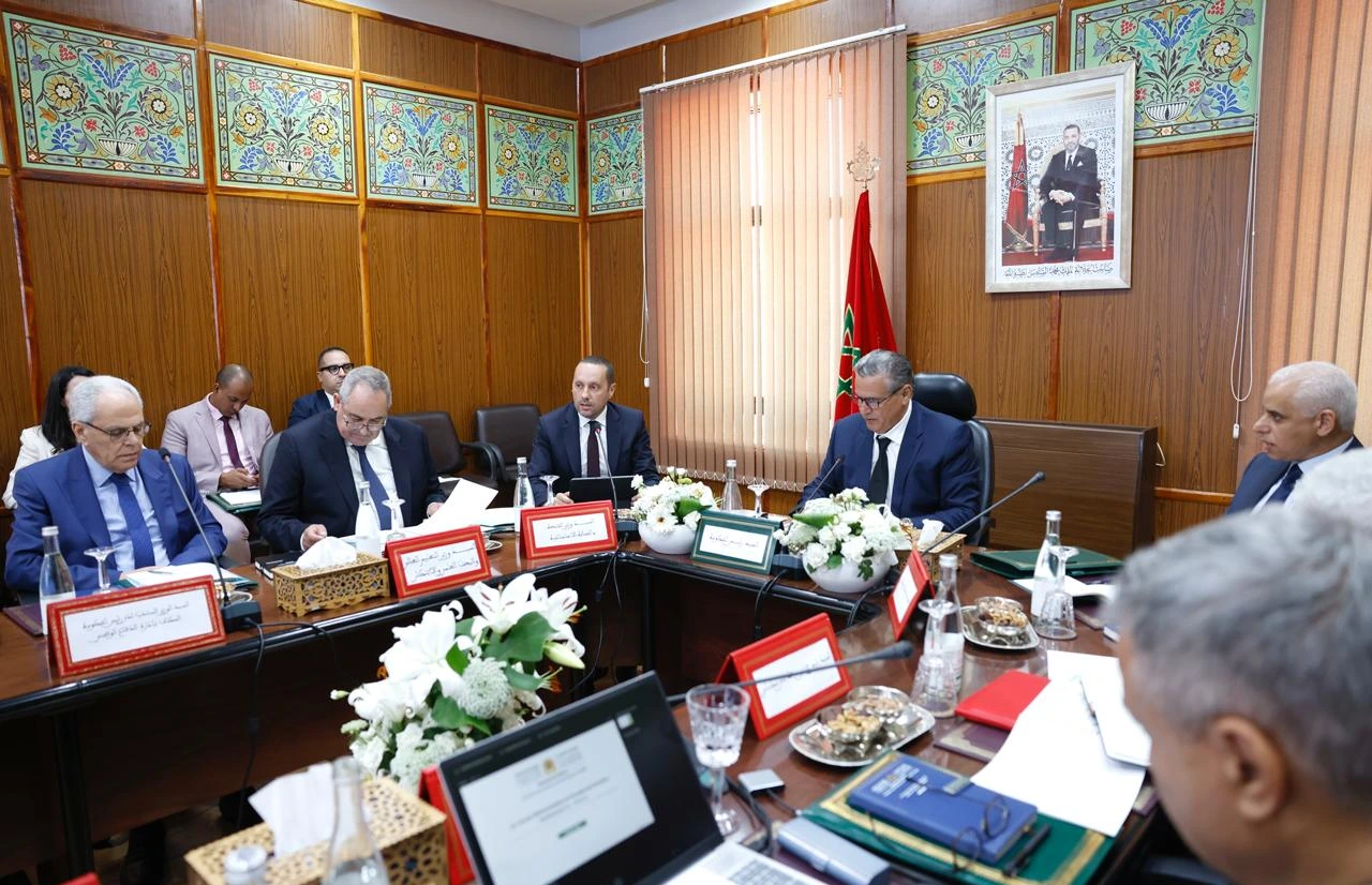 vis_0609202619525930 | MCG24 | موقع إلكتروني مغربي شامل متجدد على مدار الساعة Formal meeting around a wooden conference table with officials, Moroccan flag, and a framed portrait in an ornate wood-paneled room.
