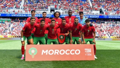 vis_0611202619583670 | MCG24 | موقع إلكتروني مغربي شامل متجدد على مدار الساعة Morocco national football team posing on the field behind a large MOROCCO banner, players in red jerseys and green shorts with fans in the stands behind them.