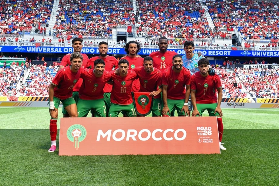 vis_0611202619583670 | MCG24 | موقع إلكتروني مغربي شامل متجدد على مدار الساعة Morocco national football team posing on the field behind a large MOROCCO banner, players in red jerseys and green shorts with fans in the stands behind them.