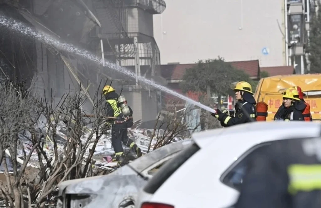 4544484 | MCG24 | موقع إلكتروني مغربي شامل متجدد على مدار الساعة Firefighters in yellow helmets spray water through smoke over a rubble-filled urban street with a damaged white car in the foreground.