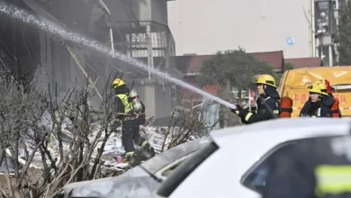 Firefighters in yellow helmets spray water through smoke over a rubble-filled urban street with a damaged white car in the foreground.