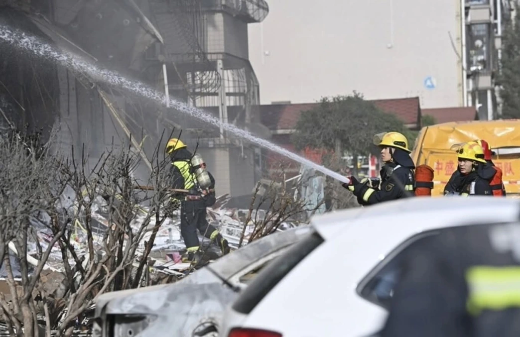 Firefighters in yellow helmets spray water through smoke over a rubble-filled urban street with a damaged white car in the foreground.