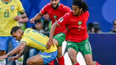 Morocco captain in a red jersey with armband dribbles past a Brazilian defender in yellow during a tense match.