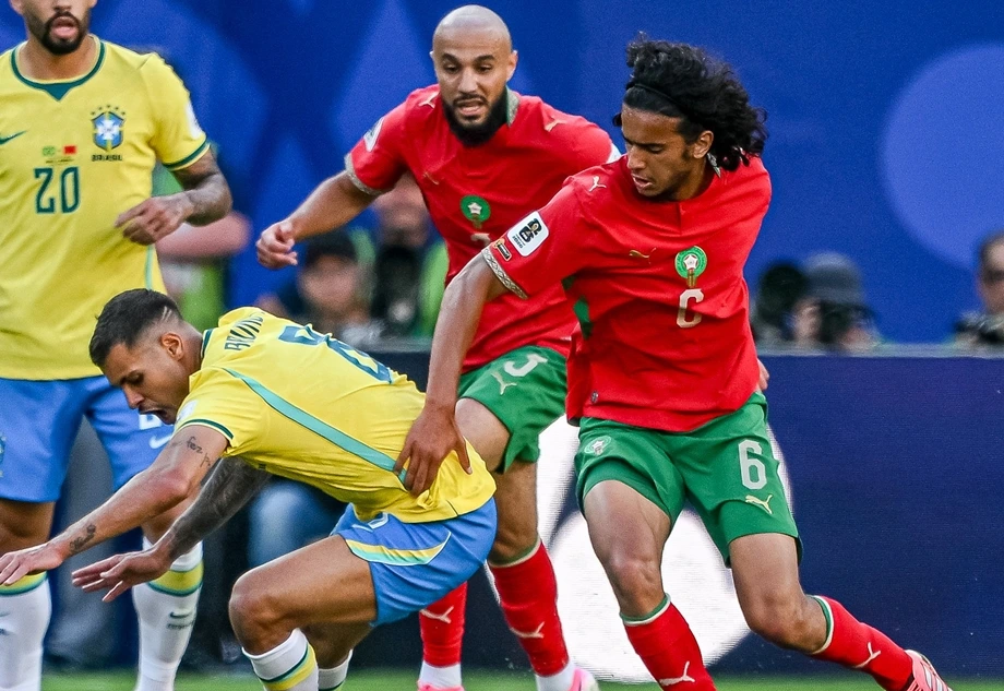 Morocco captain in a red jersey with armband dribbles past a Brazilian defender in yellow during a tense match.