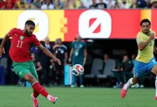 vis_0615202608382807 | MCG24 | موقع إلكتروني مغربي شامل متجدد على مدار الساعة Moroccan player in red jersey (#11) kicks a soccer ball as a Brazil defender in yellow (#4) pursues on a green field at a stadium.