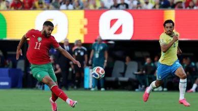 Moroccan player in red jersey (#11) kicks a soccer ball as a Brazil defender in yellow (#4) pursues on a green field at a stadium.