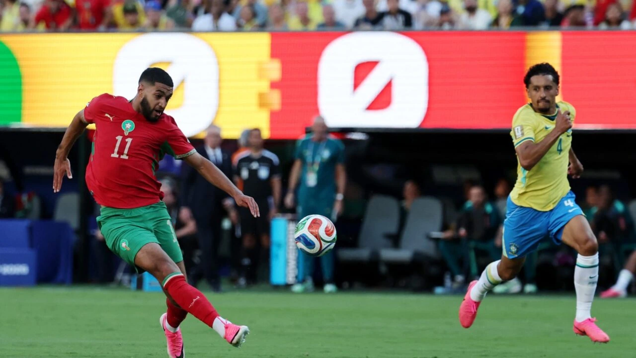 Moroccan player in red jersey (#11) kicks a soccer ball as a Brazil defender in yellow (#4) pursues on a green field at a stadium.