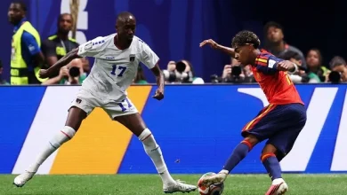 Soccer players in white and red-blue kits compete for the ball on a green field, with a defender closing in.