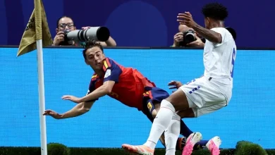 515154548 | MCG24 | موقع إلكتروني مغربي شامل متجدد على مدار الساعة Soccer scene near the corner flag with a red-and-blue jersey player sliding while a white-kit player challenges from the right as photographers watch in the background