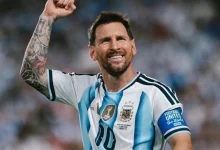 Argentine footballer celebrating on the pitch, fist raised, wearing white-and-light-blue striped jersey with the number 10 and captain armband highlight tattoos on his arm.