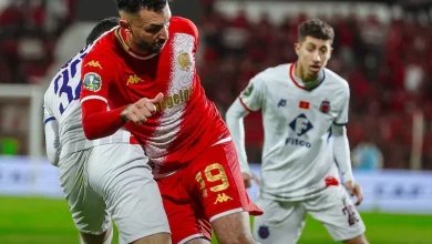 wac-ocs1 | MCG24 | موقع إلكتروني مغربي شامل متجدد على مدار الساعة Red jersey player (#19) battles for the ball with a white-jersey defender during a soccer match on a stadium field.