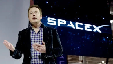 Man in a dark blazer and purple plaid shirt speaks on a stage with a SpaceX logo in the background.
