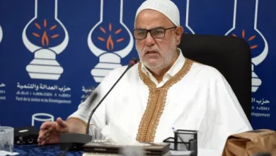 Older man wearing a white traditional robe with gold trim and a white cap speaks into a microphone at a press conference, backdrop shows blue banner with party logos and Arabic text.