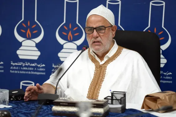 Older man wearing a white traditional robe with gold trim and a white cap speaks into a microphone at a press conference, backdrop shows blue banner with party logos and Arabic text.