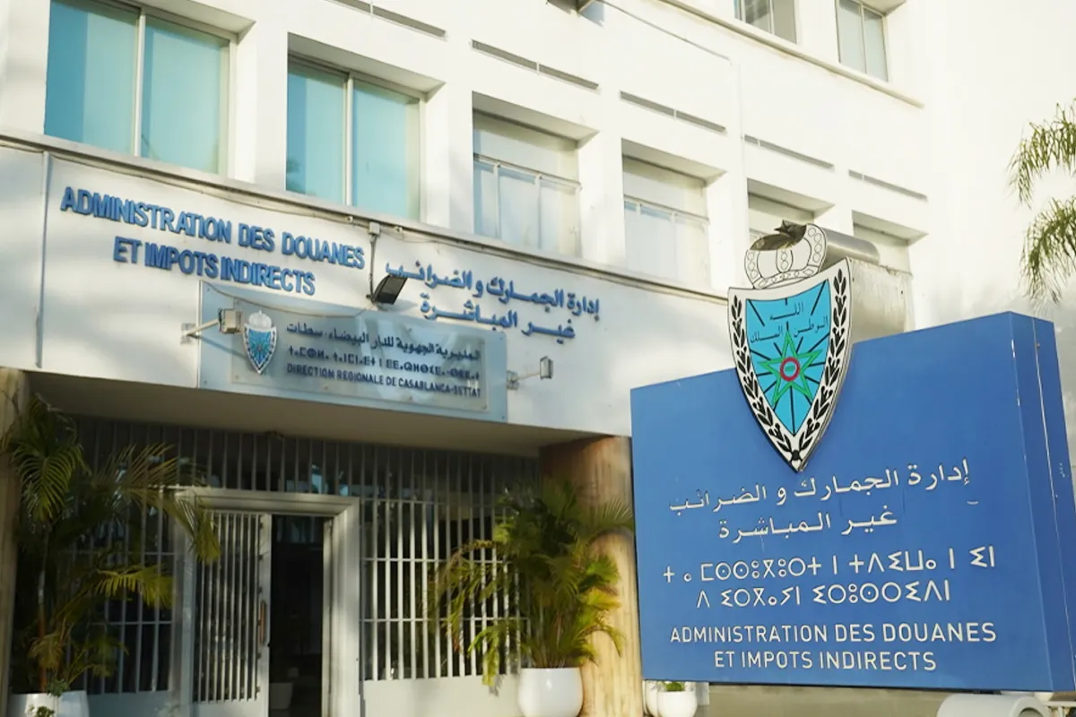 Front of a government office with blue bilingual signs and a crest, doors and white building façade behind palm plants