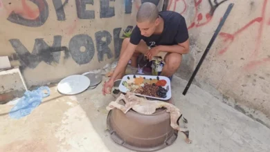 A man crouches by an outdoor clay pot stove, arranging a tray of spices while a whole animal carcass rests on the pot.