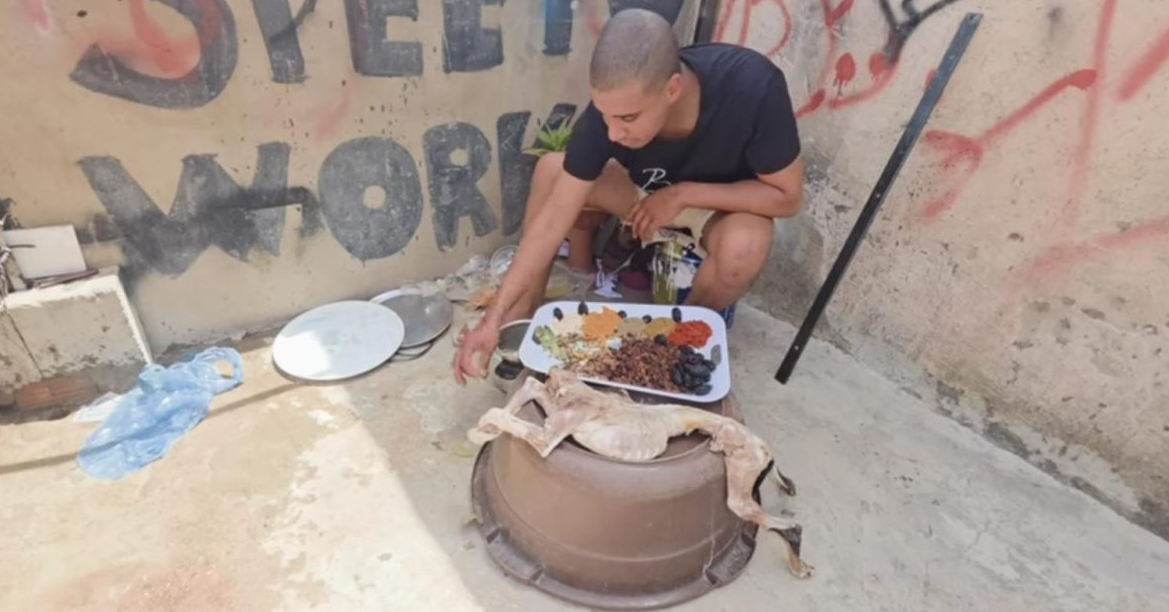 التهم المنسوبة لـ بن نسنس | MCG24 | موقع إلكتروني مغربي شامل متجدد على مدار الساعة A man crouches by an outdoor clay pot stove, arranging a tray of spices while a whole animal carcass rests on the pot.