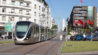 الرباطأرشيف- | MCG24 | موقع إلكتروني مغربي شامل متجدد على مدار الساعة Modern tram glides along a city street with white apartment buildings on the left and a row of red flags by a dark building on the right.