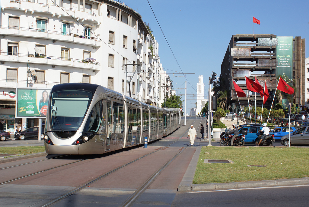 الرباطأرشيف- | MCG24 | موقع إلكتروني مغربي شامل متجدد على مدار الساعة Modern tram glides along a city street with white apartment buildings on the left and a row of red flags by a dark building on the right.