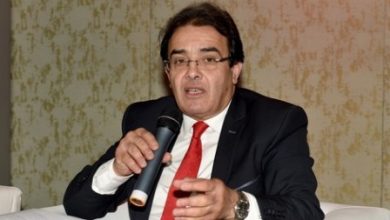 الوزير السابق عبد الكريم بنعتيق | MCG24 | موقع إلكتروني مغربي شامل متجدد على مدار الساعة Man in a dark suit and red tie speaks into a handheld microphone while seated on a white couch during a discussion.