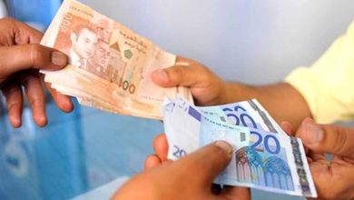 تحويلات-المغاربة-بالخارج | MCG24 | موقع إلكتروني مغربي شامل متجدد على مدار الساعة Two people exchanging cash, one handing over a stack of brownish banknotes while the other receives a bundle of blue and purple notes across a table.