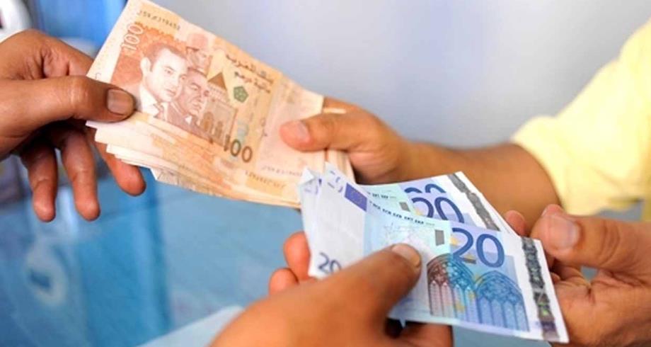 تحويلات-المغاربة-بالخارج | MCG24 | موقع إلكتروني مغربي شامل متجدد على مدار الساعة Two people exchanging cash, one handing over a stack of brownish banknotes while the other receives a bundle of blue and purple notes across a table.