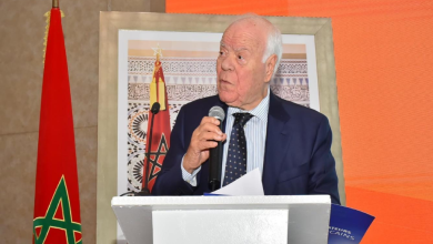 حسن السنتيسي | MCG24 | موقع إلكتروني مغربي شامل متجدد على مدار الساعة Older man in a dark blue suit speaks into a microphone at a white podium, with a Moroccan flag and framed artwork behind him.