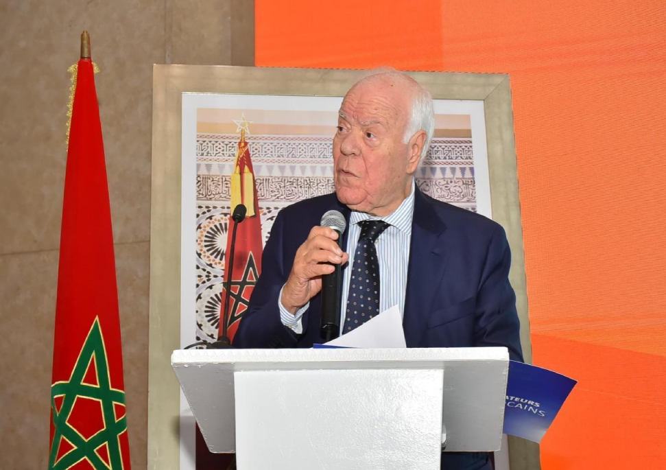 حسن السنتيسي | MCG24 | موقع إلكتروني مغربي شامل متجدد على مدار الساعة Older man in a dark blue suit speaks into a microphone at a white podium, with a Moroccan flag and framed artwork behind him.