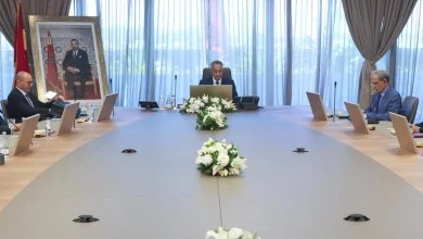 Formal meeting room with a long conference table, officials seated on both sides, and a floral centerpiece with a framed portrait on the left wall.