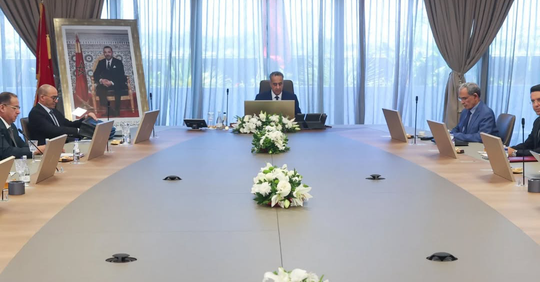 Formal meeting room with a long conference table, officials seated on both sides, and a floral centerpiece with a framed portrait on the left wall.