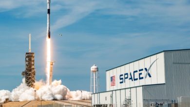 SpaceX rocket lifting off from a launch pad beside a large SpaceX building, with smoke and a bright flame plume behind it.