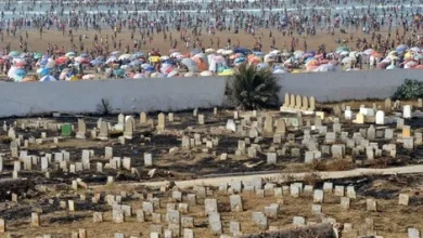 Overhead view of a crowded beach with colorful umbrellas along the shore, and a dirt cemetery with many white gravestones in the foreground separated by a wall.