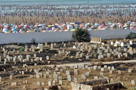 شاطئ الرباط | MCG24 | موقع إلكتروني مغربي شامل متجدد على مدار الساعة Overhead view of a crowded beach with colorful umbrellas along the shore, and a dirt cemetery with many white gravestones in the foreground separated by a wall.