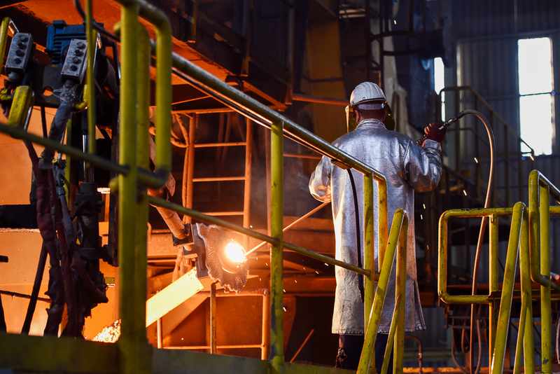 Worker in protective gear welds metal on an industrial line; sparks fly near orange-lit machinery and yellow safety rails.