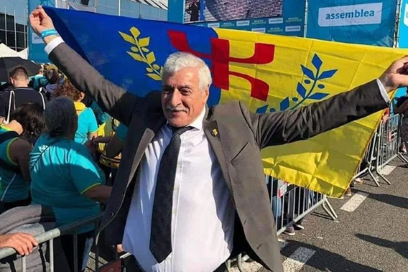 Older man in a suit with arms raised in celebration at a political rally, yellow and blue banner behind him-to the left and right.