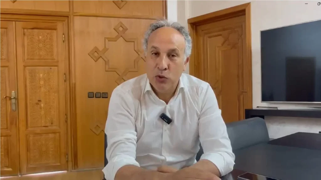Man in a white shirt seated at a table in a living room with wooden paneled doors behind him.