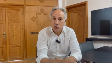 كودارjpg | MCG24 | موقع إلكتروني مغربي شامل متجدد على مدار الساعة Man in a white shirt seated at a table in a living room with wooden paneled doors behind him.