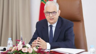 Man in a dark suit and glasses sits at a conference table with documents and a floral centerpiece, speaking in a formal meeting.”,