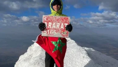 Climber on a snowy summit holding a sign that reads 'ARARAT 5165 m' with a Moroccan flag draped over their torso.