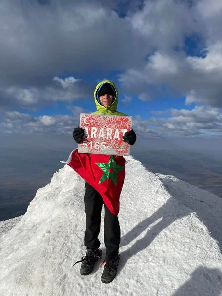 Climber on a snowy summit holding a sign that reads 'ARARAT 5165 m' with a Moroccan flag draped over their torso.
